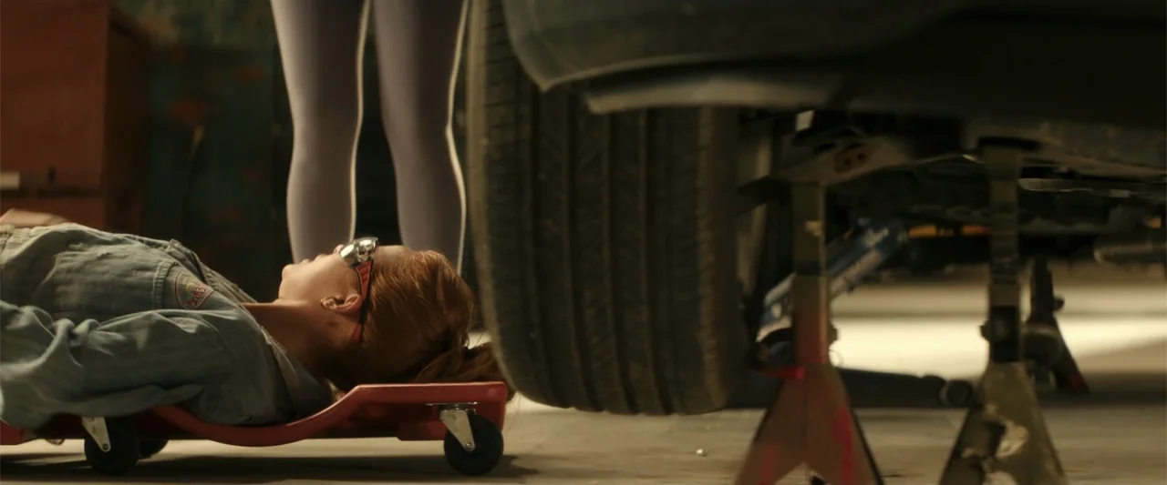Maintenance Required (2025) movie still: medium — An auto repair shop interior contains a car raised on metal jack stands. A woman lies on a red…