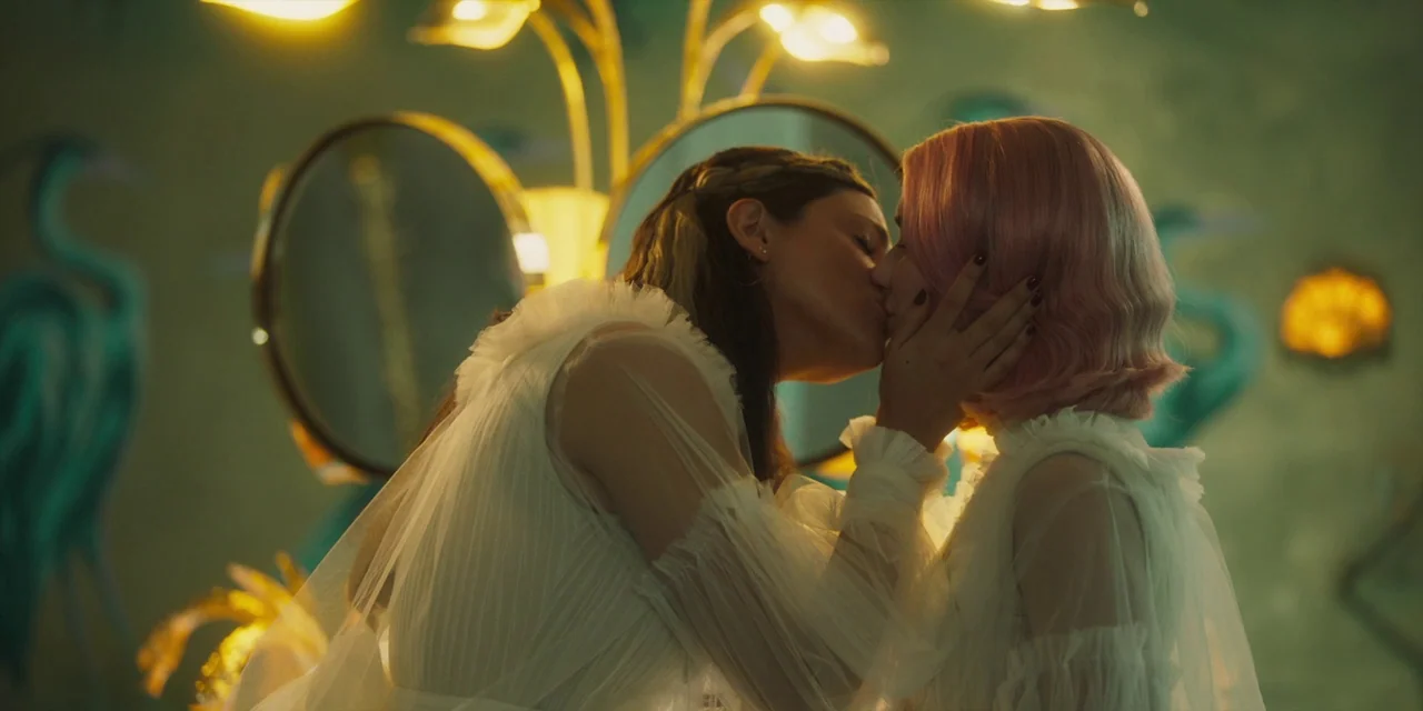 Paradise Hills (2019) movie still: medium — Two women stand in front of illuminated oval mirrors in a room with green walls. They are kissing while…