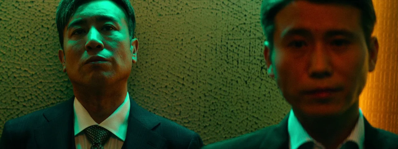 Under The Light (2023) movie still: medium — Two men in dark business suits stand closely together against a textured wall surface. They remain…
