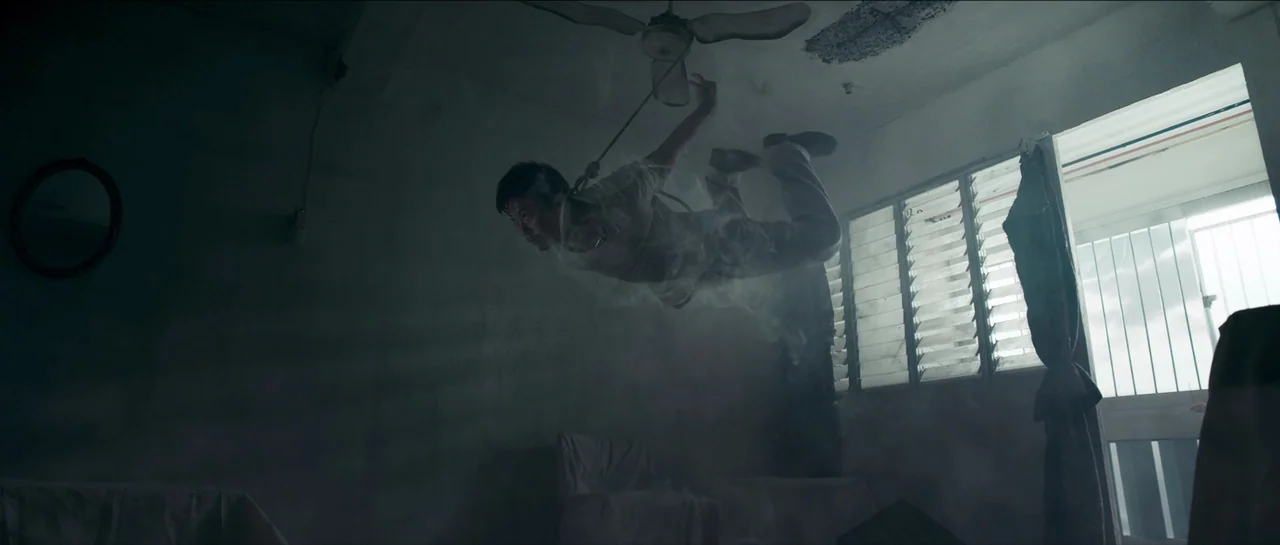 Rigor Mortis (2013) movie still: fullbody — A dimly lit room with peeling paint features a man suspended by a ceiling fan. Dense particles of dust…