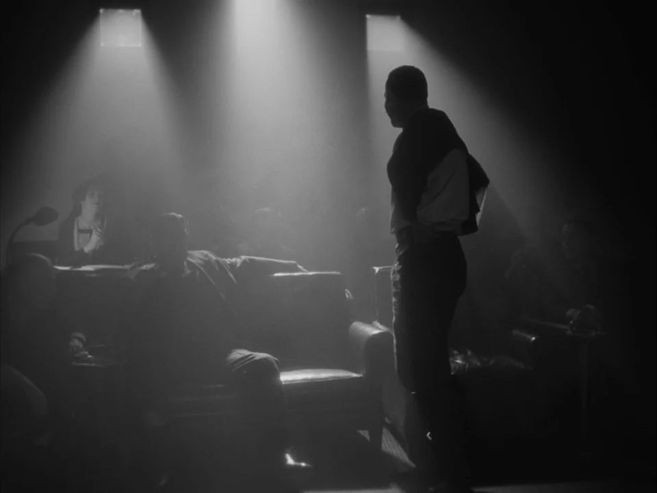 The Artist (2011) movie still: medium — A dimly lit room with multiple people seated on leather armchairs. One man stands facing a seated man amidst…