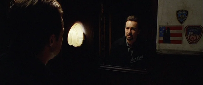 25th Hour (2002) movie still: medium — A dimly lit restroom with a wall-mounted sconce between the viewer and the mirror. The man looks directly at…