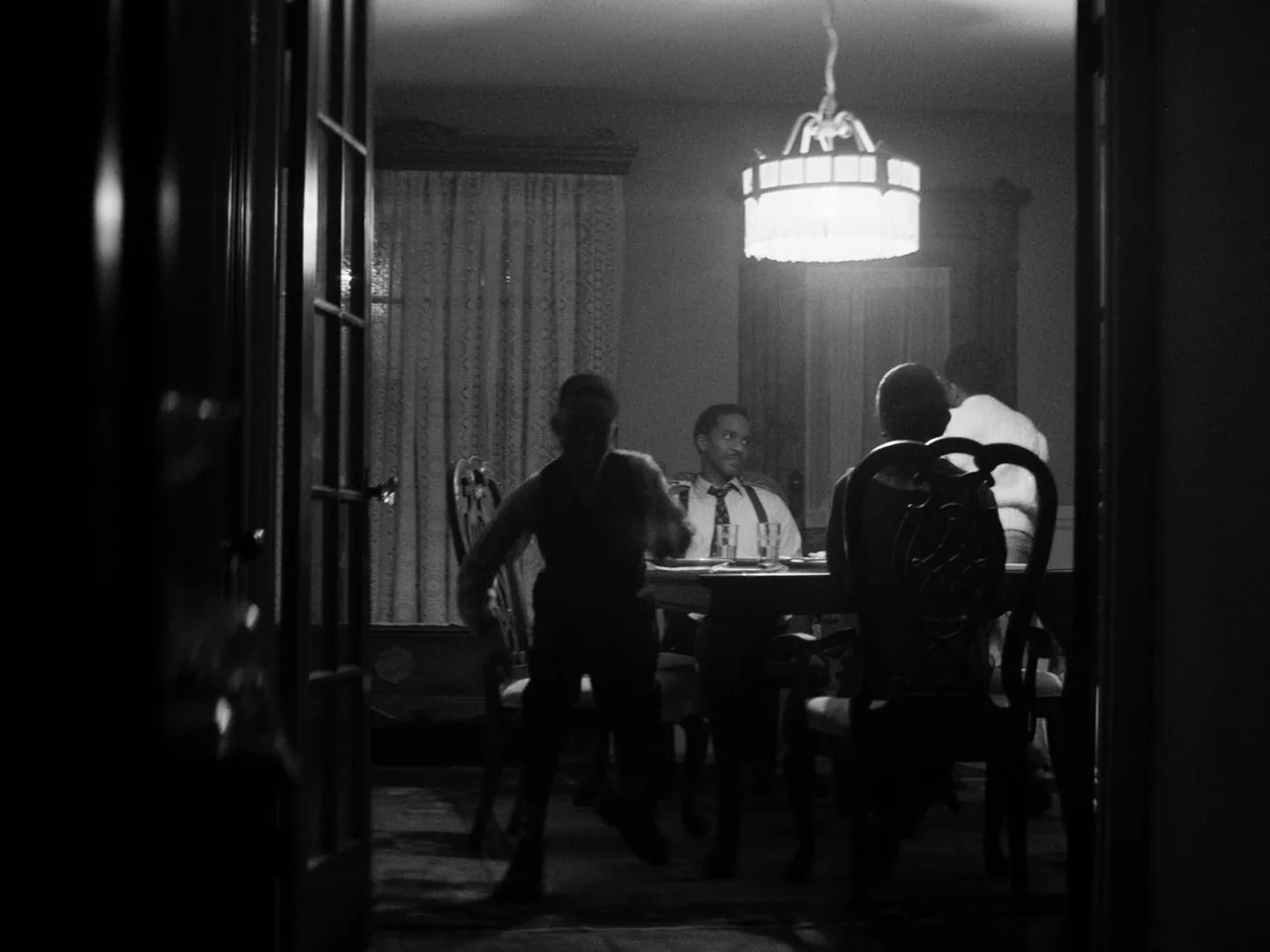Passing (2021) movie still: medium — A dimly lit room with an ornate wooden table and heavy curtains in background. An adult man sits at the head of…