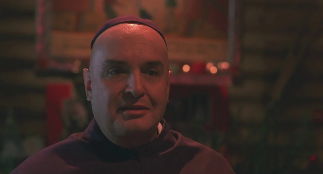 Santa Sangre (1989) movie still: closeup — A dimly lit interior space with a religious painting faintly visible on the wall. A middle-aged man in…