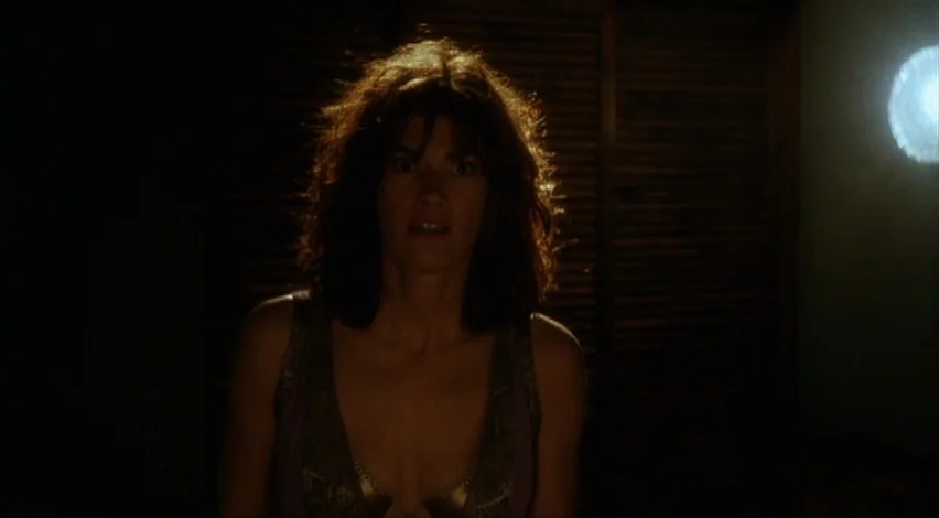 Dust Devil (1992) movie still: medium — A dimly lit room features a dark louvered panel in the background. A woman stands in the foreground, facing…