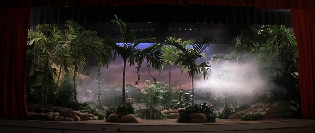 Rushmore (1998) movie still: establishing — A theatrical stage features an elaborate jungle set with artificial palm trees and mounds. Thick…