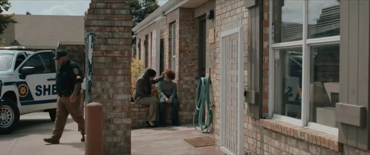 Possessions (2024) movie still: wide — A brick building exterior with a white gate and a sheriff vehicle parked nearby. A woman rests her hand on…