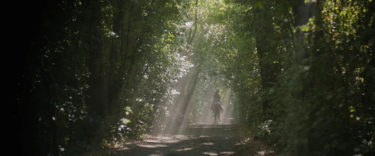 Rosaline (2022) movie still: wide — A dirt path cuts through a dense forest with thick overhanging foliage. A rider on a horse travels down the…