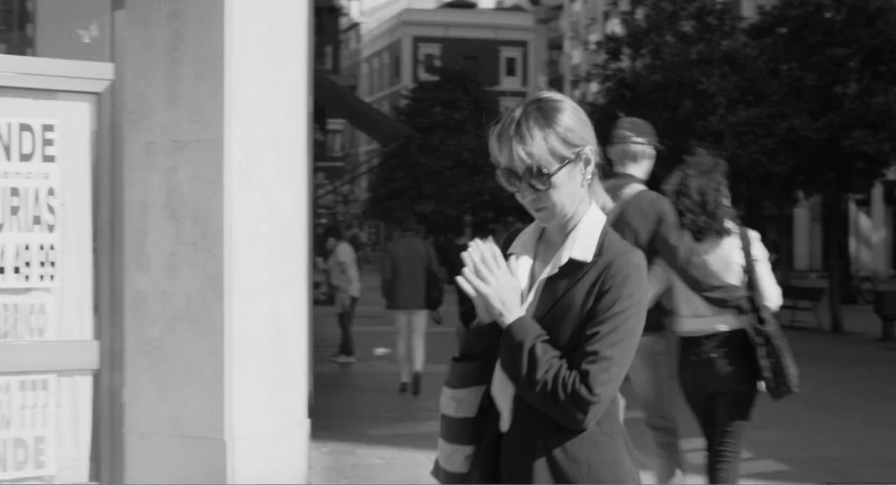 El Planeta (2021) movie still: medium — A sunny city sidewalk features pedestrians moving past a building facade with posted signage. A woman…