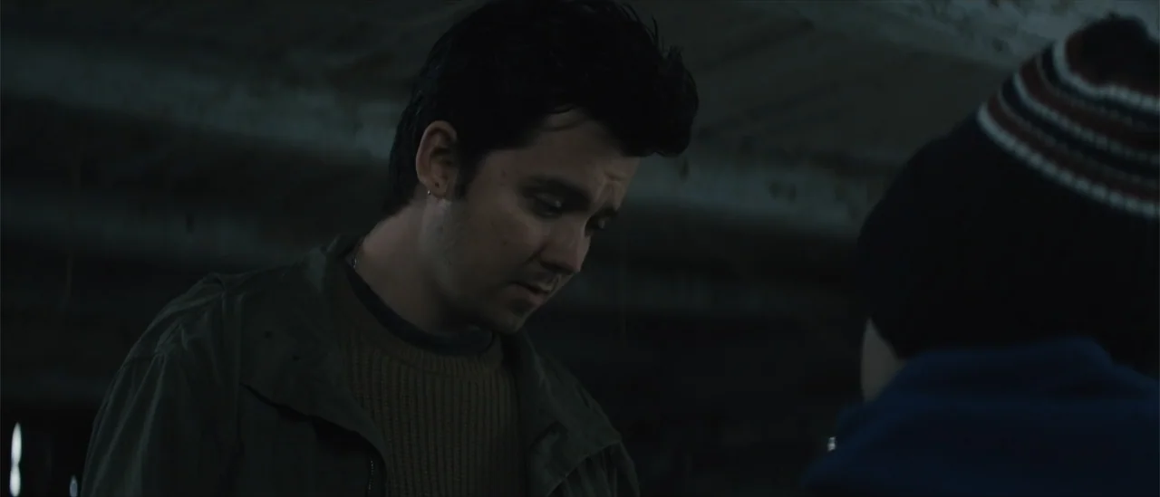 All Fun and Games (2023) movie still: medium — A dark concrete basement with indistinct textures on the ceiling and walls. A young man in a brown…