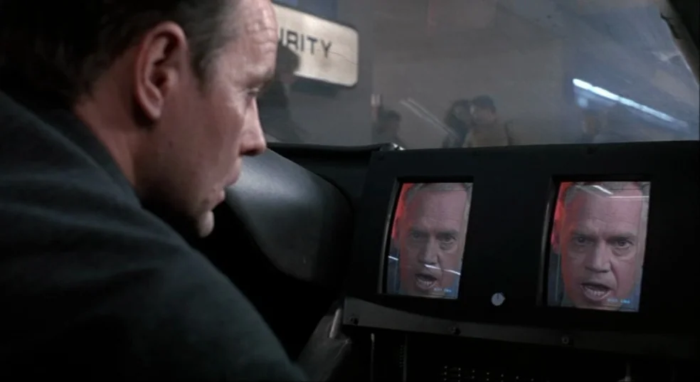 Total Recall (1990) movie still: medium — The interior of a vehicle features a dashboard with two small embedded video screens. A man in the…