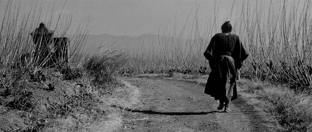 Yojimbo (1961) movie still: wide — A desolate rural landscape features a narrow dirt path bordered by tall leafless shrubs. A lone man wearing…
