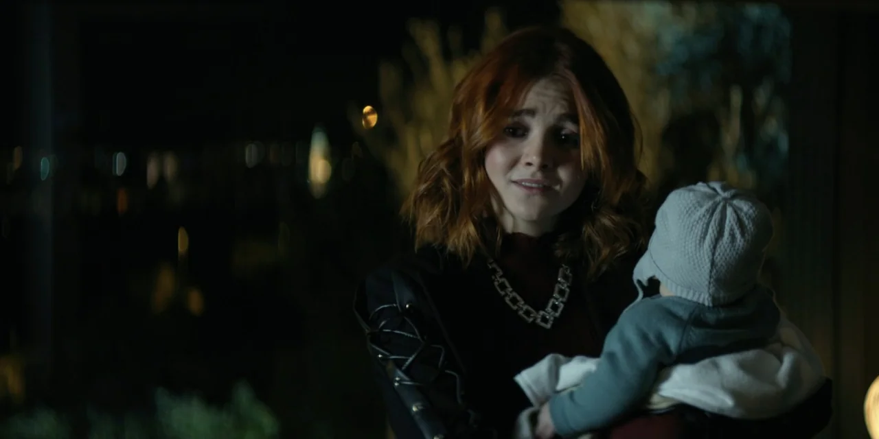 Our Fault (2025) movie still: medium — A dark outdoor space at night with blurred light bokeh in the background. A woman with auburn hair holds a…