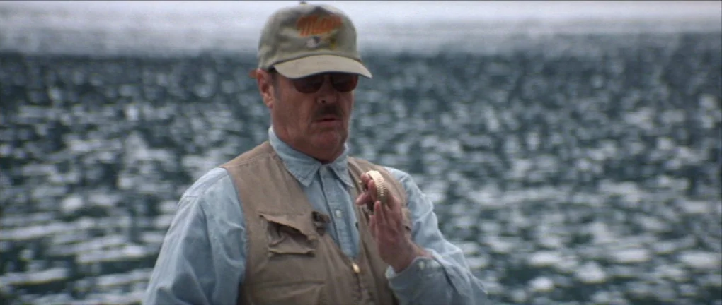 The Pledge (2001) movie still: medium — A man stands outdoors near a body of water with a choppy, grey surface. He wears a fishing vest and cap…