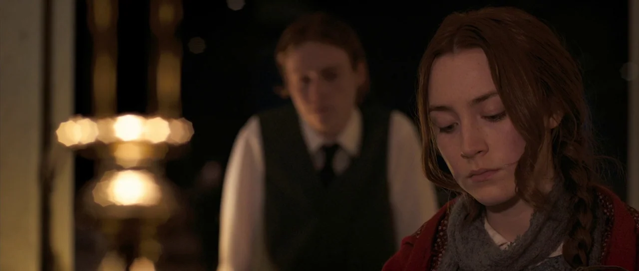 Byzantium (2012) movie still: medium — A dimly lit indoor space with dark tones and a blurred background. A woman sits in the foreground with a man…