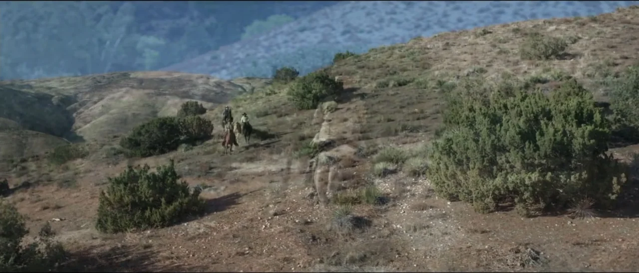 Dead for a Dollar (2022) movie still: wide — An arid hillside with sparse vegetation and scrub brush under a clear blue sky. Two individuals on…