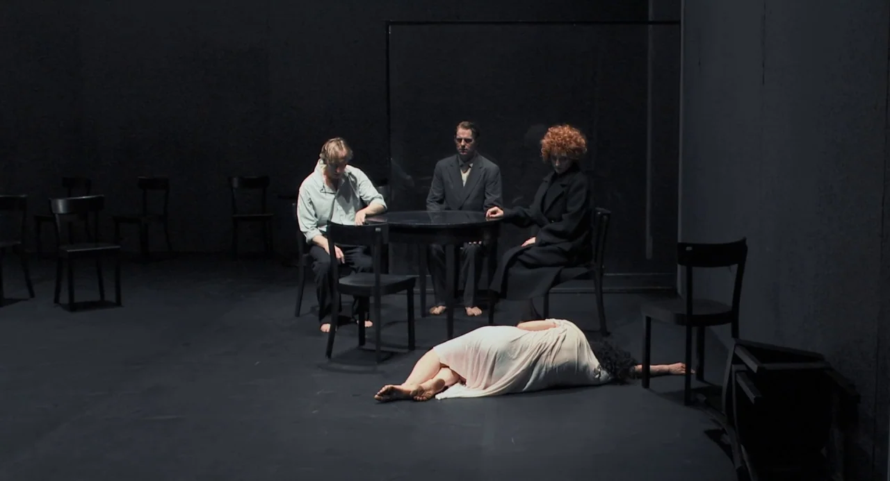 Pina (2011) movie still: wide — A dark, empty theatrical space contains only a small round table and several wooden chairs. Three people sit at the…
