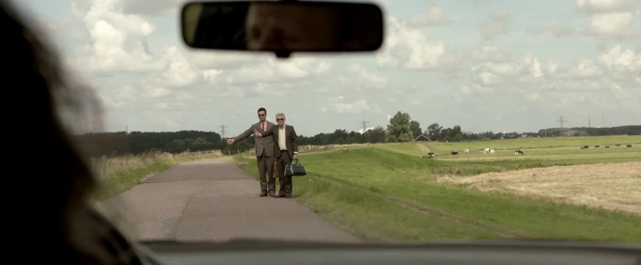 Borgman (2013) movie still: wide — A quiet rural road cuts through a green pastoral landscape with distant grazing cattle. Two men in formal…