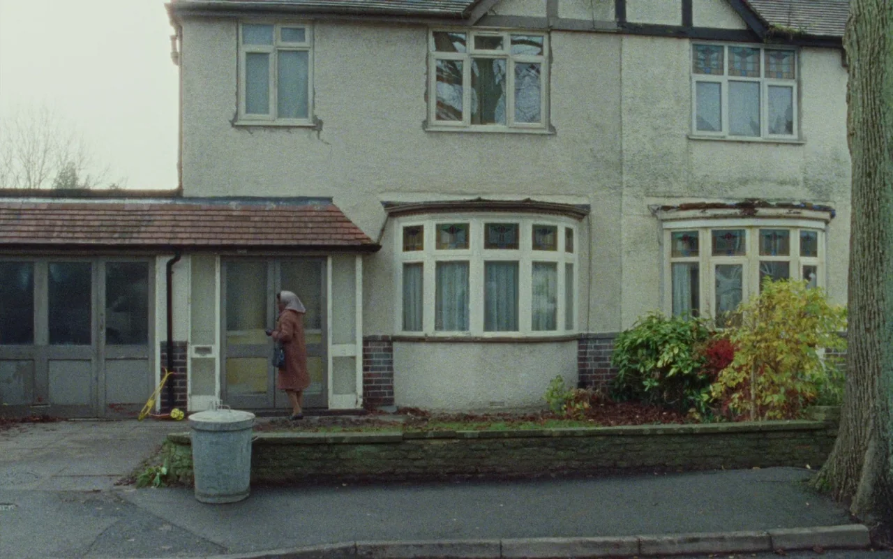 Education (2020) movie still: wide — A two-story suburban house features light-colored render and multiple bay windows. A woman stands by the side…