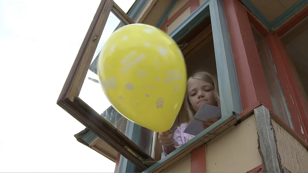 Boathouse Detectives (2010) movie still: medium — A girl looks out from an open wooden window frame of a building. She holds a bright yellow…