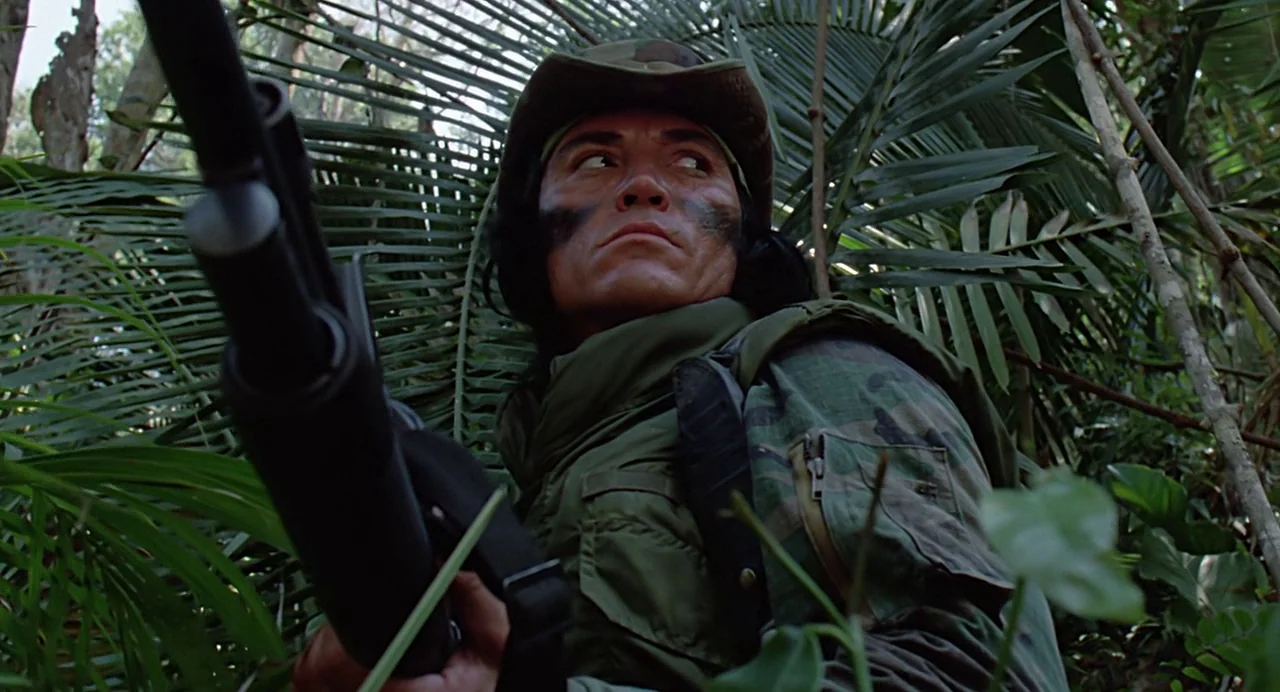 Predator (1987) movie still: medium — A dense, humid jungle environment filled with lush green palms and tangled vegetation. A soldier in camouflage…