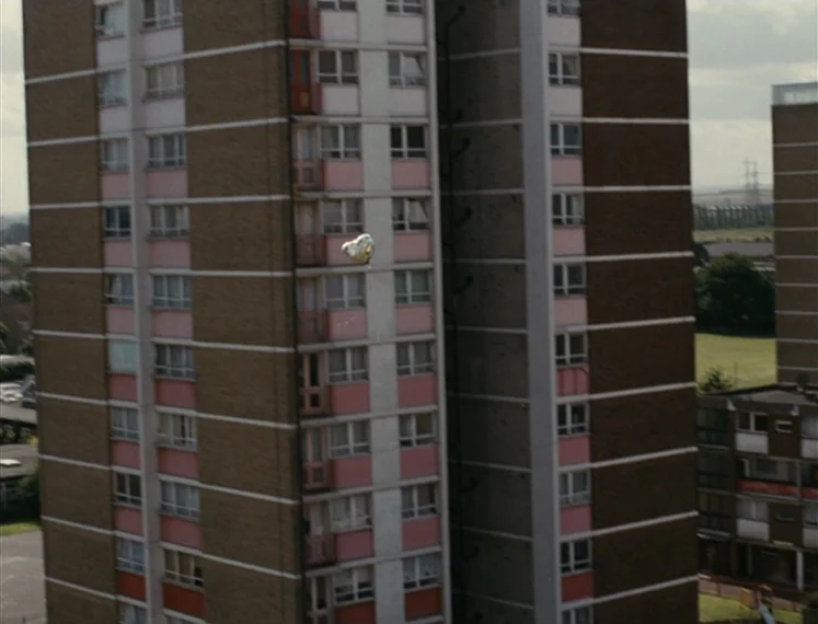 Fish Tank (2009) movie still: establishing — A tall mid-century concrete and brick apartment tower sits against a cloudy sky. A small reflective…