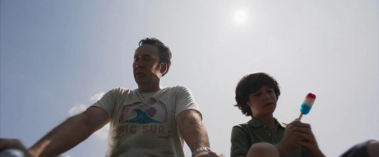 Mom and Dad (2017) movie still: medium — A man and a young boy are positioned outdoors against a clear, bright sky. The man wears a Big Sur graphic…