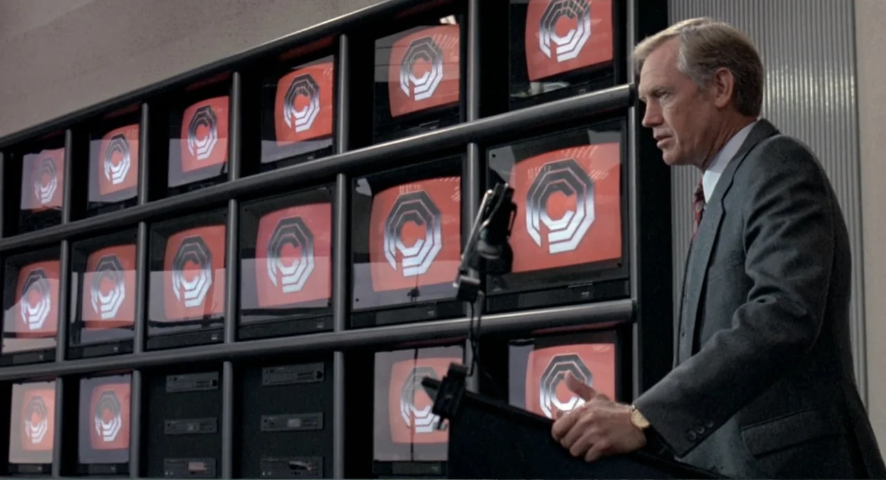 RoboCop (2014) movie still: medium — A man in a suit stands at a podium in front of a video wall. Multiple monitors display a red and white…