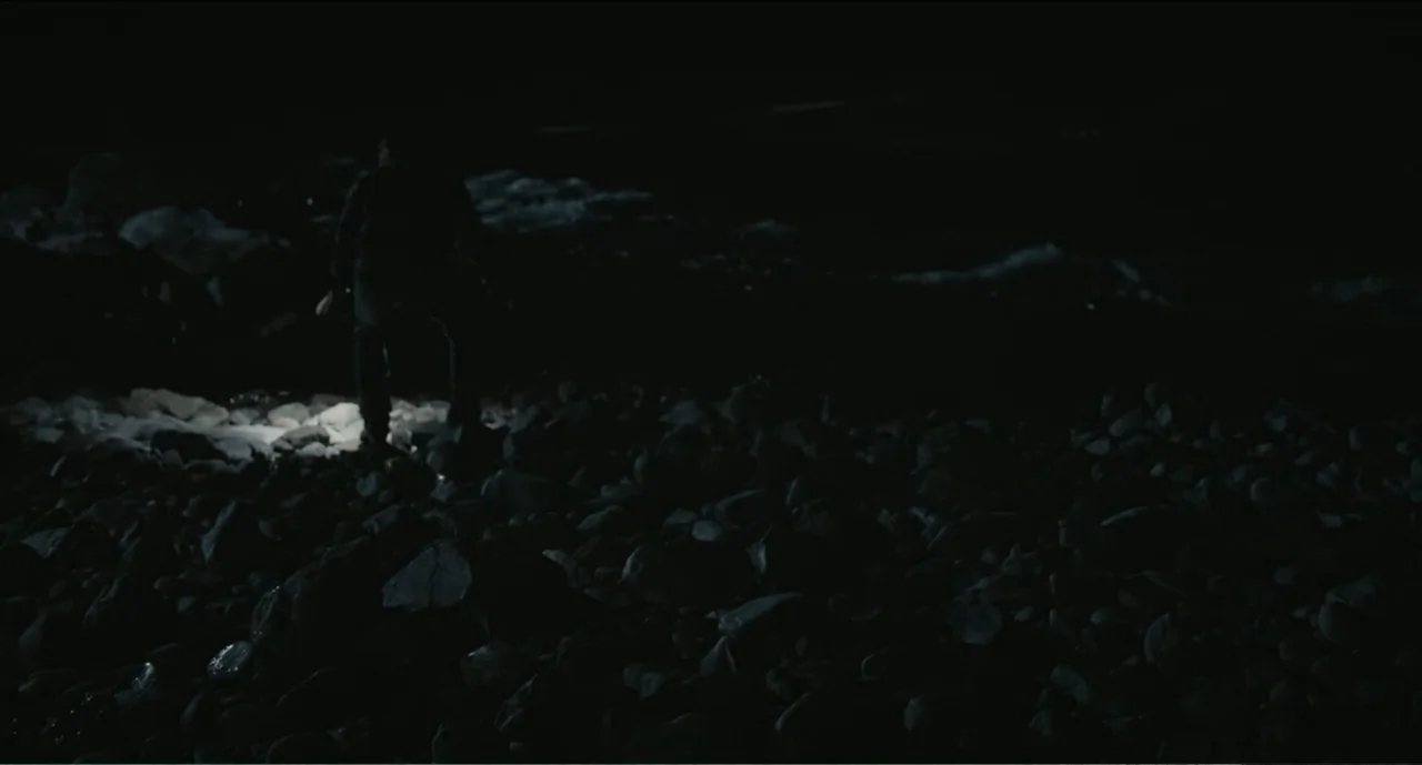 Cowboys (2014) movie still: wide — A dark, rocky shoreline at night stretches across the frame. A single person stands amidst the stones illuminated…