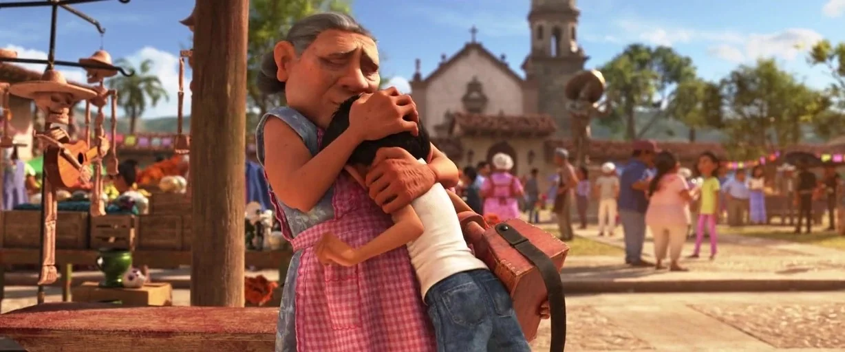 Coco (2017) movie still: medium — A sunny town square with a church visible in the background and merchant stalls nearby. An older woman hugs a…