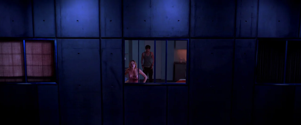 Kaboom (2010) movie still: wide — A flat concrete wall with three rectangular windows separates a dark hallway from a room. A blonde woman and a man…