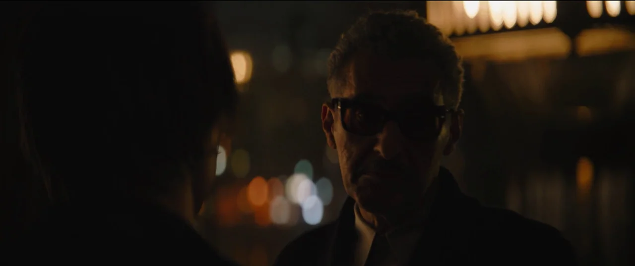 The Batman (2022) movie still: medium — A city street at night with out-of-focus bokeh lights in the dark background. An older man in dark glasses…