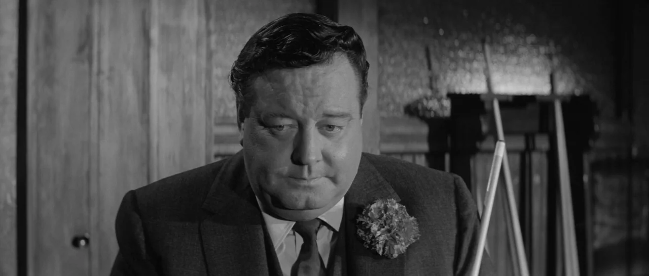 The Hustler (1961) movie still: closeup — A dimly lit room features a man standing in front of wall-mounted cues. The subject is wearing a suit with…