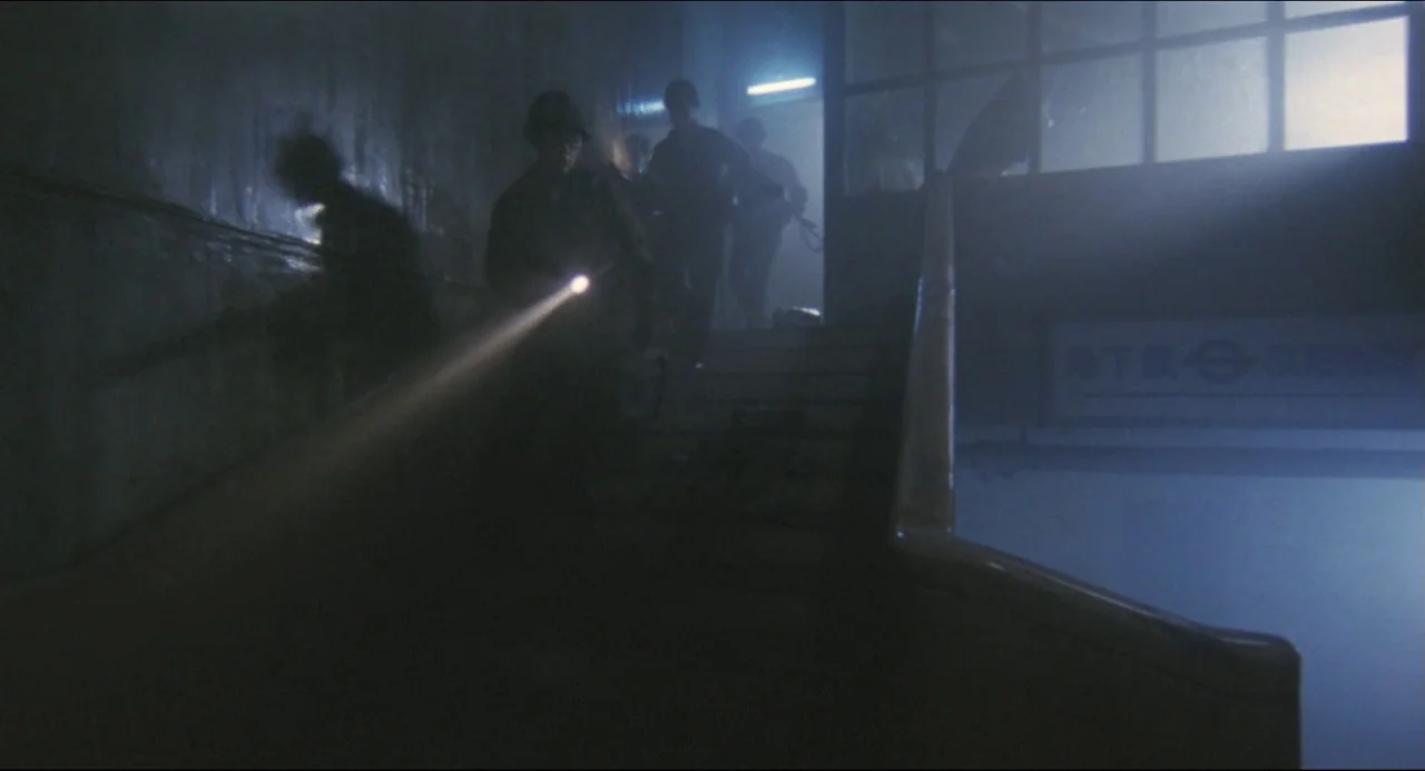 Godzilla vs. Biollante (1989) movie still: medium — A dimly lit subway entrance with hazy air and concrete walls. Three soldiers in tactical gear…