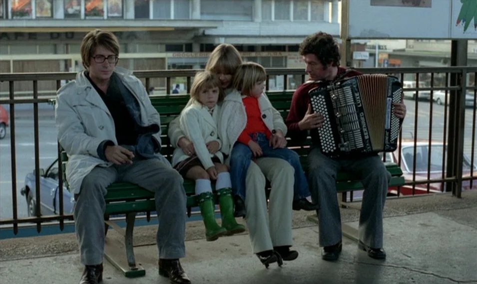 Every Man for Himself (1980) movie still: fullbody — A public outdoor platform with a metal railing overlooks a parking area with buildings. Five…