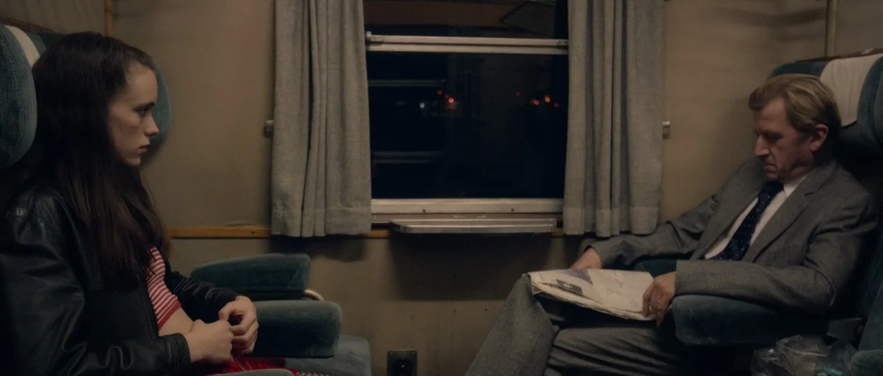 Nymphomaniac Vol 1 (2013) movie still: medium — An interior train carriage features blue upholstered seats and a dark window at night. A young woman…