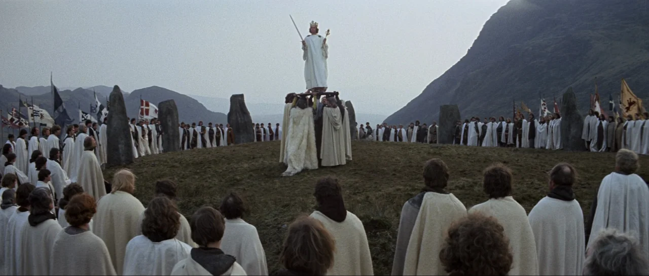 Macbeth (1971) movie still: wide — A grassy hill encircled by tall monoliths and people wearing cream colored cloaks. A king stands elevated on a…