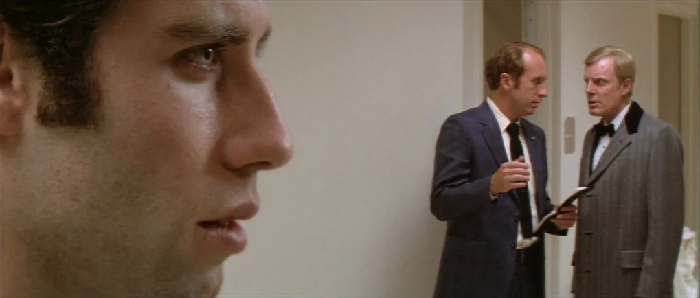 Blow Out (1981) movie still: medium — A man in the foreground remains out of focus while two men talk nearby. The setting is a brightly lit,…