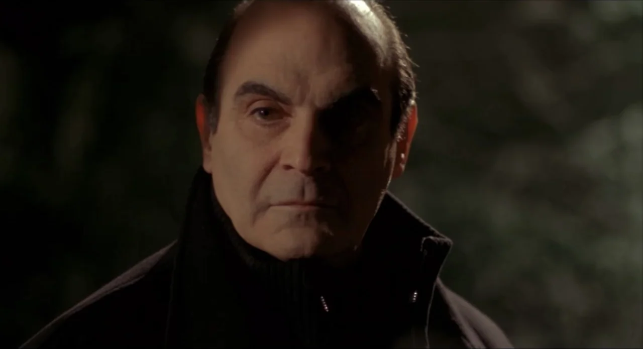 Foolproof (2003) movie still: closeup — An older man is centered in the frame against a dark, indistinct backdrop. The subject looks directly into…