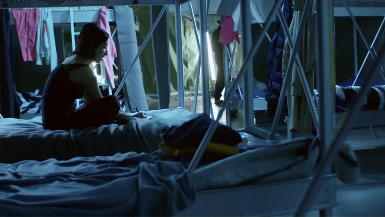 Code of Trust (2019) movie still: medium — A cluttered dormitory room contains rows of white metal bunk beds with grey blankets. A young woman sits…