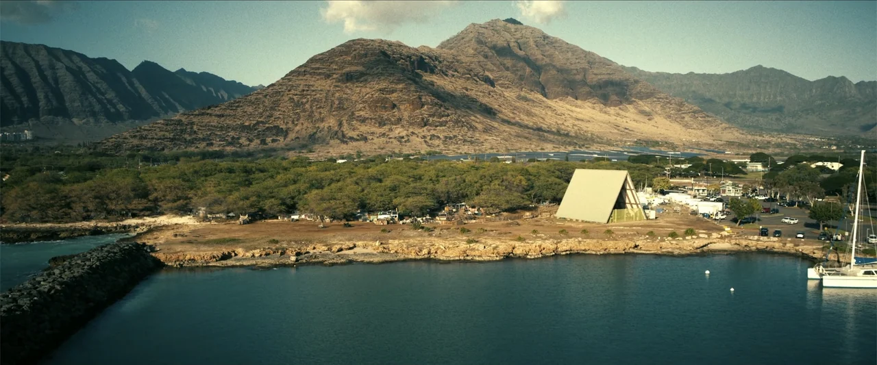The Wrecking Crew (2026) movie still: establishing — A rugged coastline sits beneath towering arid mountains with a calm blue harbor foreground. A…
