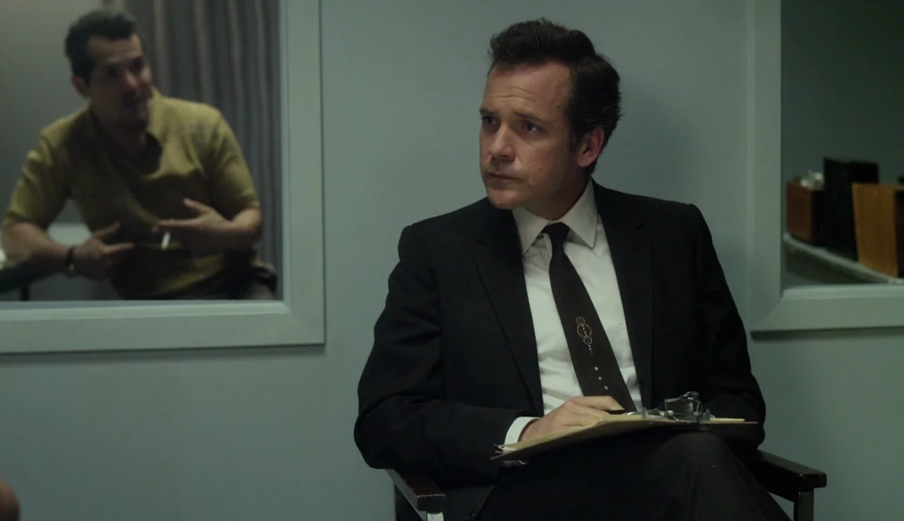 Experimenter (2015) movie still: medium — A man in a dark suit sits in a stark room with gray walls. Behind him, another man in a yellow shirt is…
