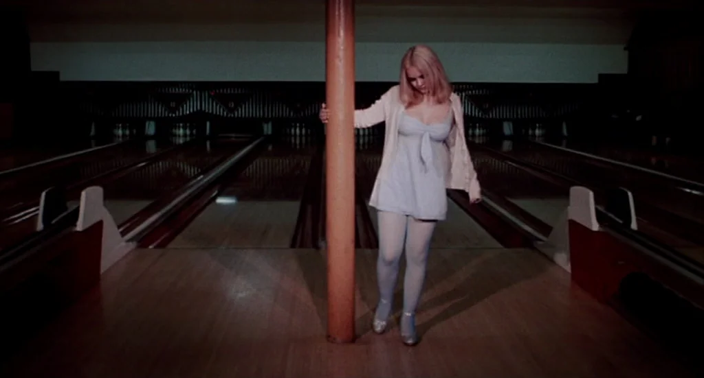 Buffalo ’66 (1998) movie still: fullbody — A dimly lit bowling alley features dark polished lanes and a prominent wooden support post. A young woman…