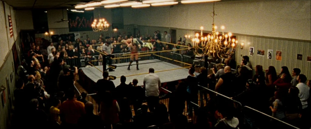 The Wrestler (2008) movie still: wide — A crowded community center hall hosts a small-scale professional wrestling match under chandeliers. A ring…