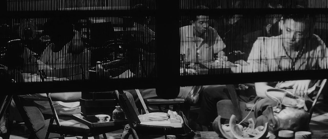 High and Low (1963) movie still: medium — Two men eat at a table viewed through a vertical wooden slatted room divider. The table is filled with…
