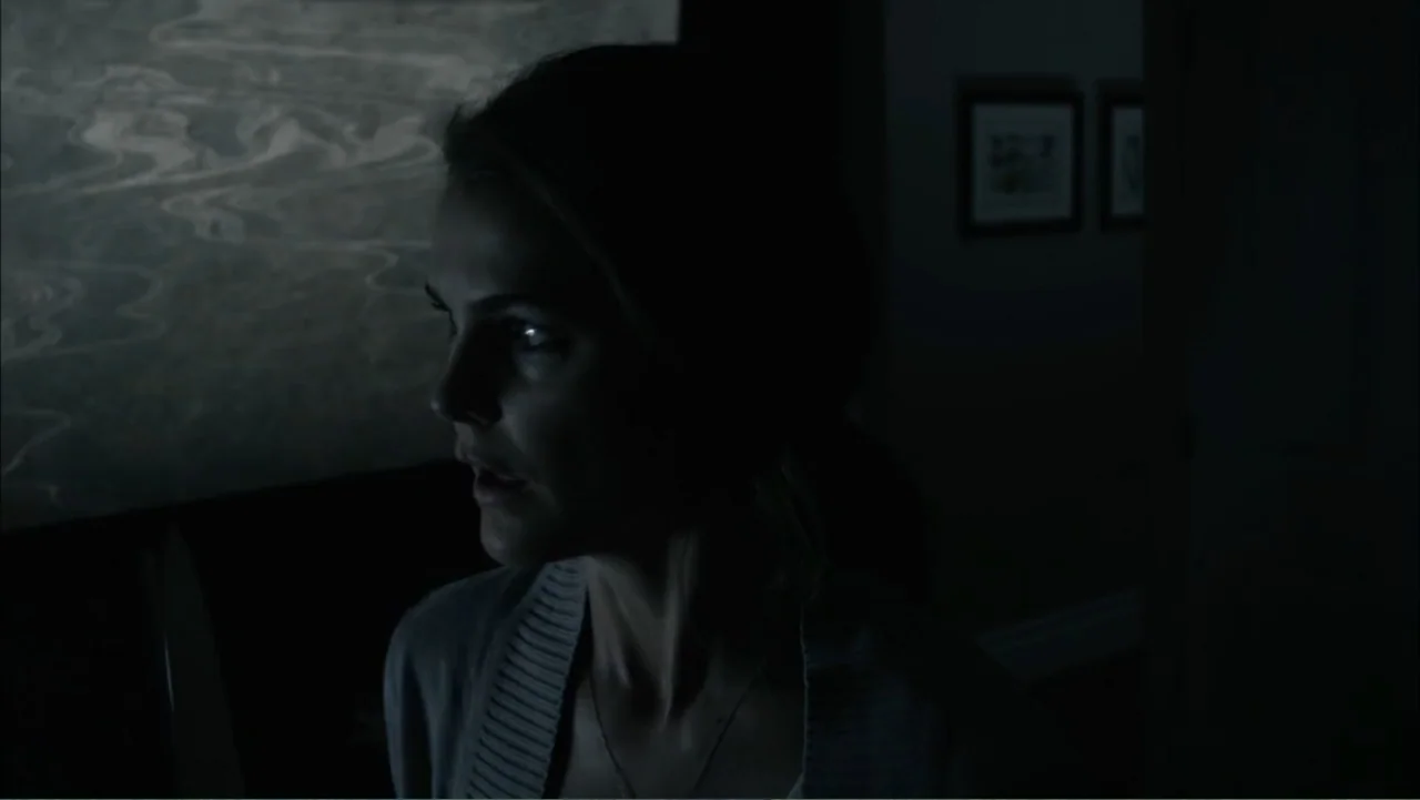 Dark Skies (2013) movie still: closeup — A dimly lit room features a woman partially illuminated against a dark background. The woman turns her head…