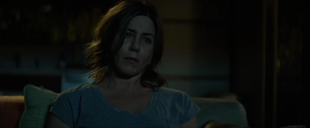 Cake (2014) movie still: medium — A dark living room with muted color tones and soft shadows. A middle-aged woman sits on a sofa looking toward the…