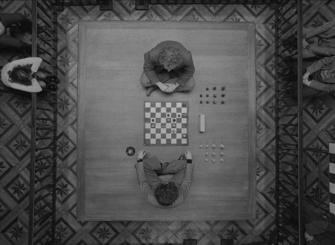 The French Dispatch (2021) movie still: wide — A high-angle view of a patterned floor where two men play checkers. Others sit observing around the…