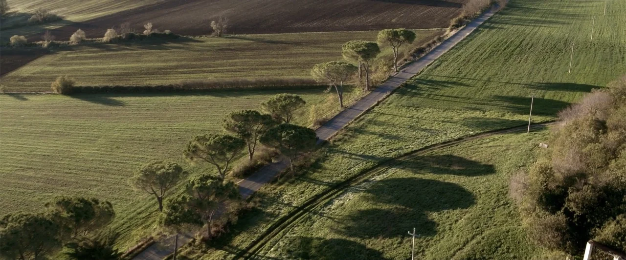 About Us (2017) movie still: establishing — A rural asphalt road winds through rolling green agricultural fields in the countryside. The scene…