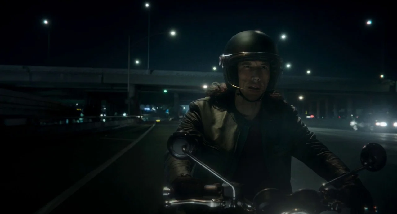 Annette (2021) movie still: medium — A man rides a motorcycle along a multilane highway illuminated by distant streetlamps. Concrete overpasses and…